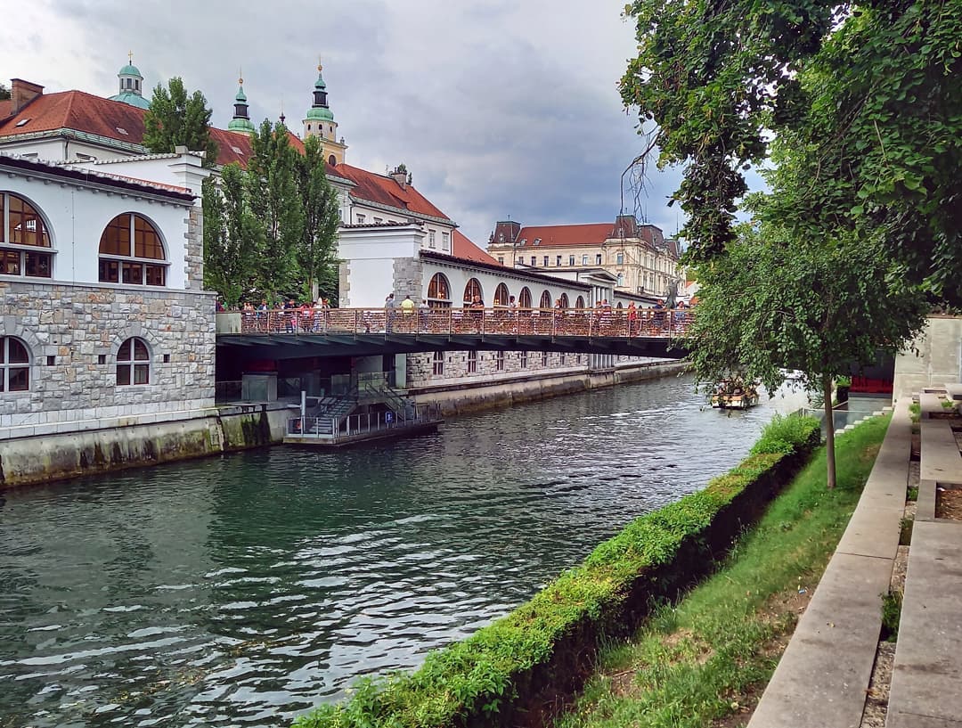 Argine del fiume di Lubiana: un ponte stretto lo attraversa, una barchetta in lontanza. Sull'altro argine si vedono i portici del mercato coperto